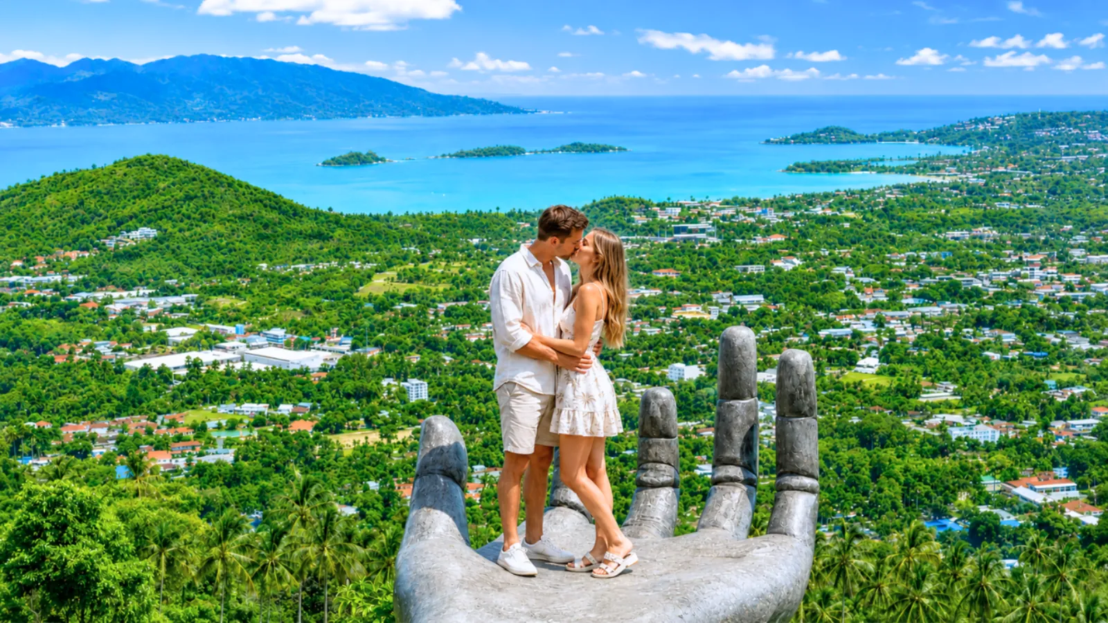A couple standing on the Big Hand sculpture at the Samui viewpoint above the coast