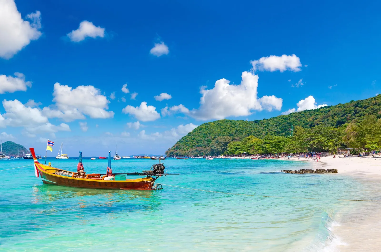 A longtail boat anchored off a beach in clear turquoise water below a wooded headland